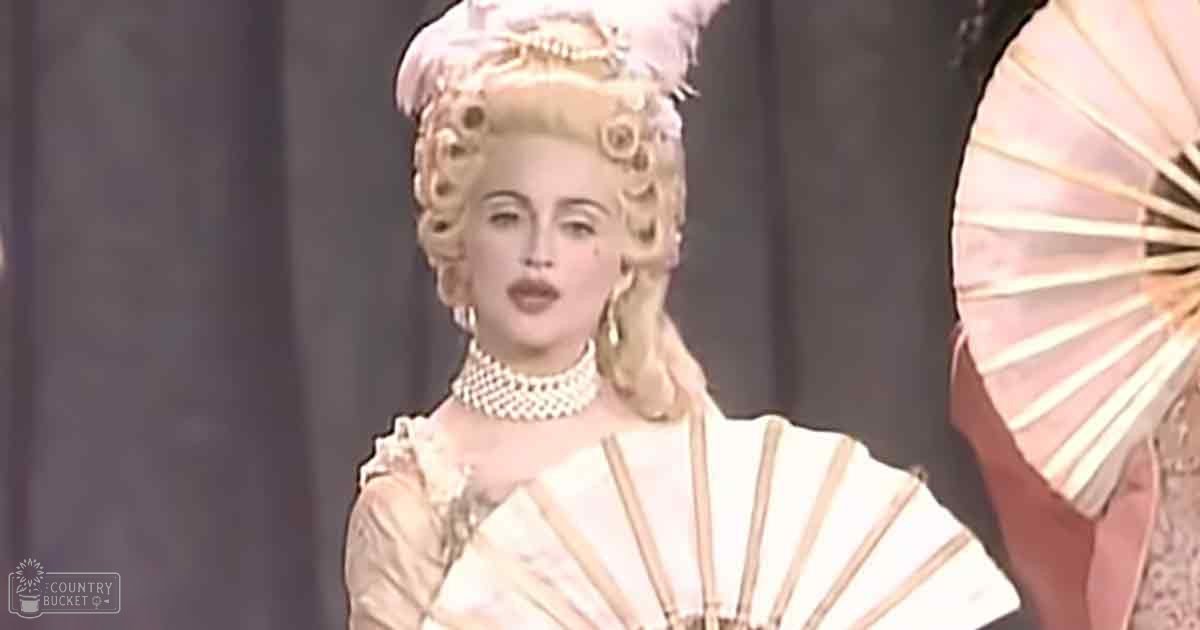 A female performer in a white and gold ornate dress, possibly from the Baroque era, holding a large white fan on stage with curtains as background. The setting suggests an indoor performance at night.