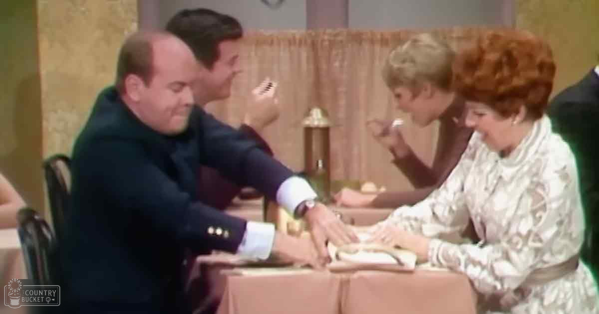 A formal indoor event with a man in a dark blue suit and women in light dresses, surrounded by furniture and a bottle on a table.