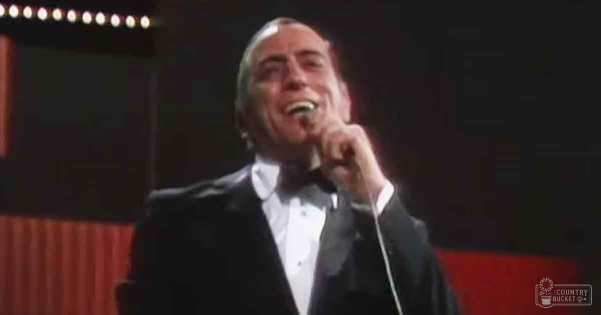 An adult male singer in a black tuxedo with white shirt and bow tie holds a microphone on stage under red curtains, suggesting a performance setting.