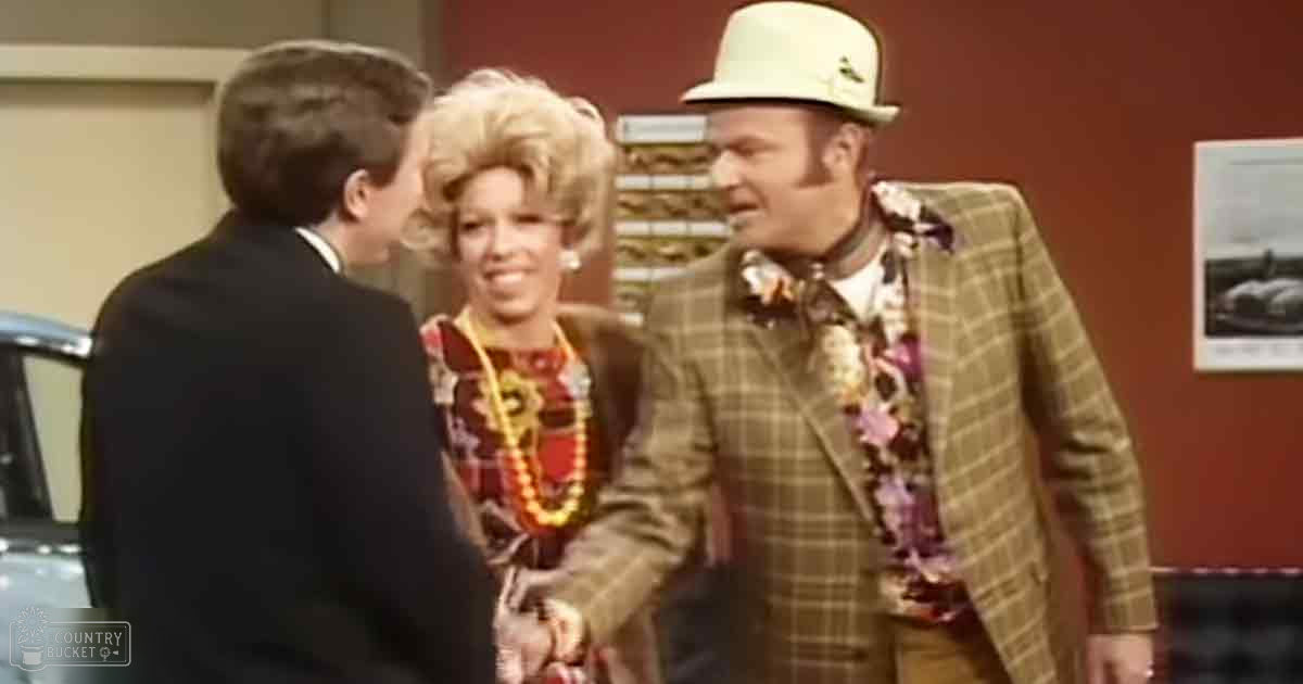 A woman in her adult years wearing a patterned floral dress indoors, accessorized with multiple necklaces and earrings; a middle-aged man in the foreground dressed in a plaid blazer over a colorful patterned tie and white hat; an adult male in the background wearing a dark suit jacket.