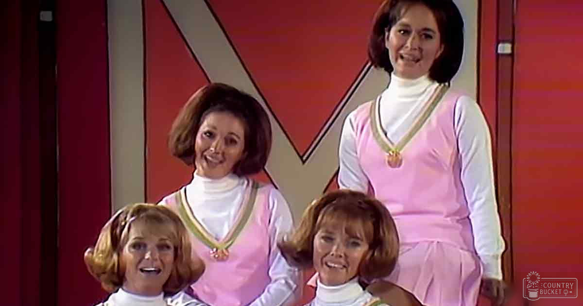 A group of four adult female performers in a mid-20th-century style setting with vibrant pink dresses and white turtlenecks, standing indoors against a red 'X' backdrop.