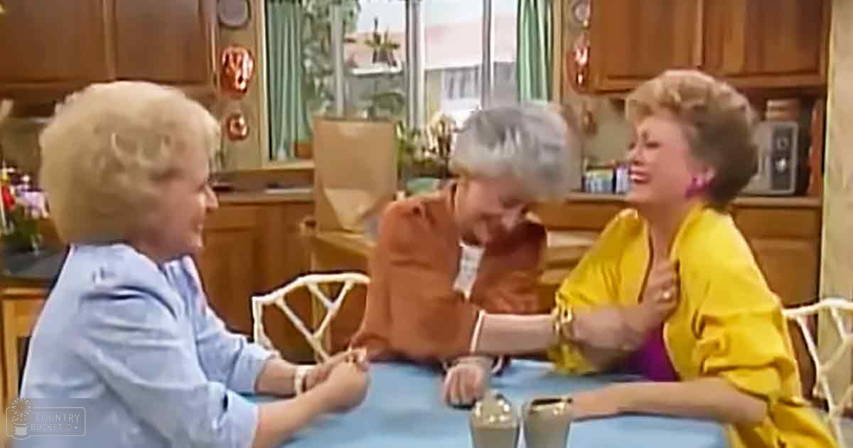 A social interaction in a kitchen setting with three older adults wearing casual jackets and accessories, wooden cabinetry, green curtains, natural daylight, blue tablecloth.