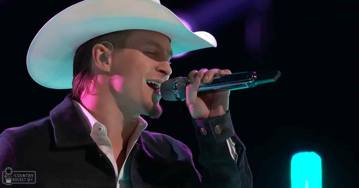 A young adult male wearing a white cowboy hat, dark jacket with buttons, and unbuttoned white shirt on stage at an indoor concert venue under purple and blue stage lights.