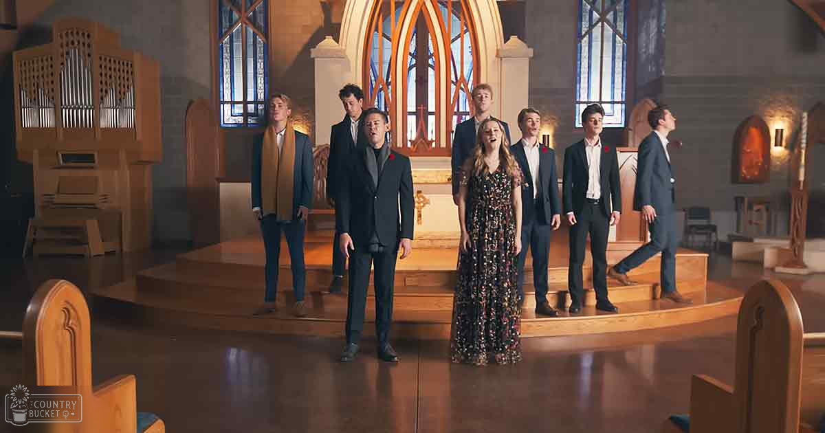 A choir of teenagers and a young adult in formal attire performing inside a church with stained glass windows allowing natural light to filter through.