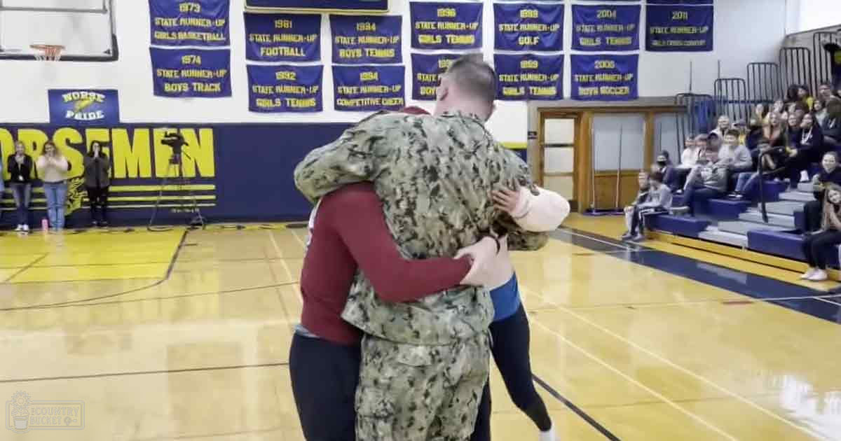 A soldier hugging a child in a gymnasium with banners listing sports achievements on the wall.