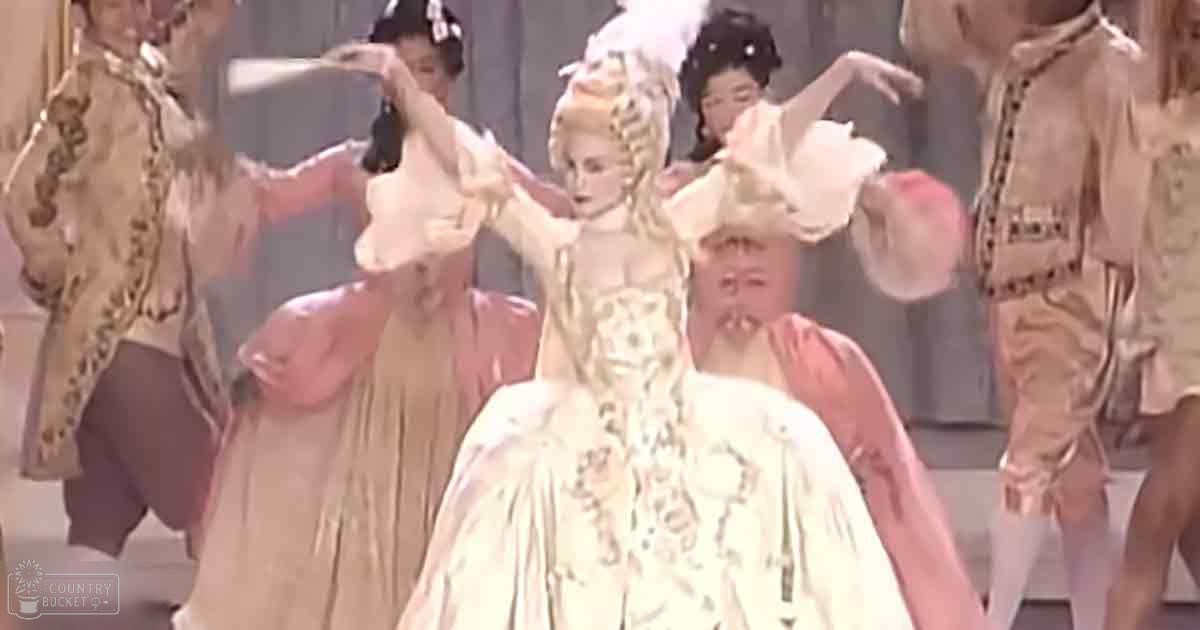 Theatrical performance scene with multiple performers in ornate costumes, set against a formal stage backdrop.