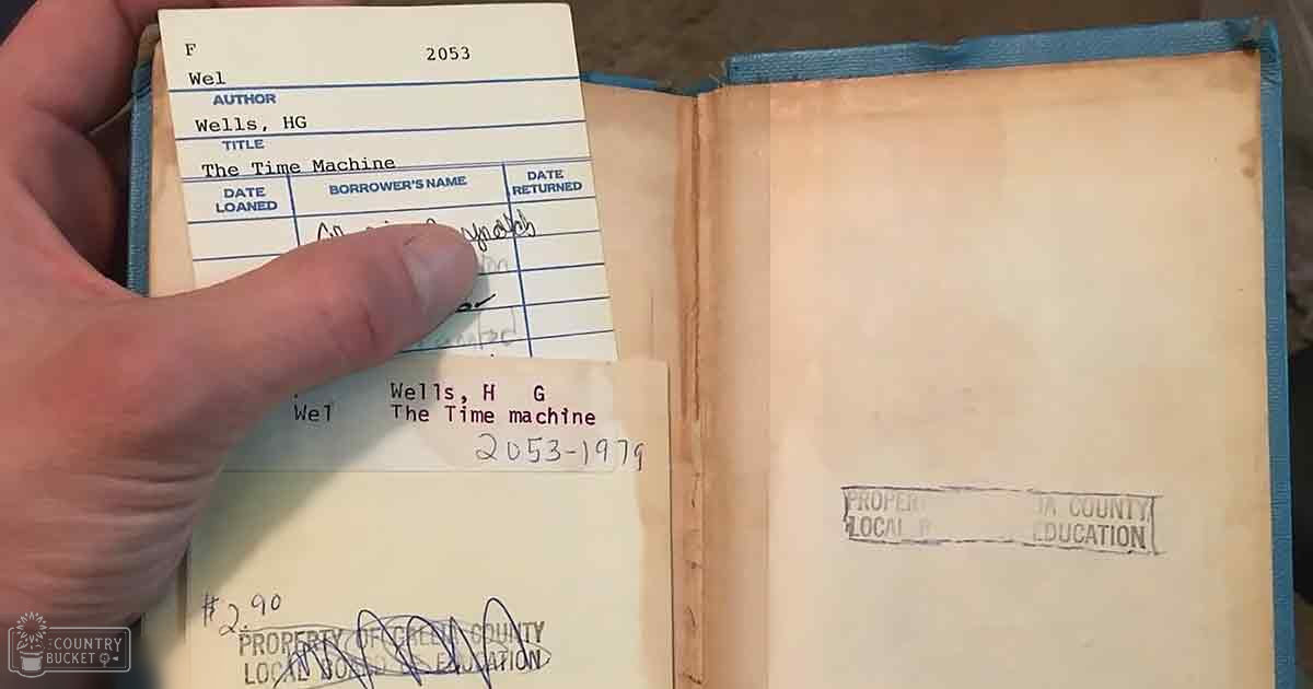 An open book titled 'The Time Machine' by H.G. Wells, with a library card inserted into it; located in Creek County Local School Education, indoor setting, natural light, daytime