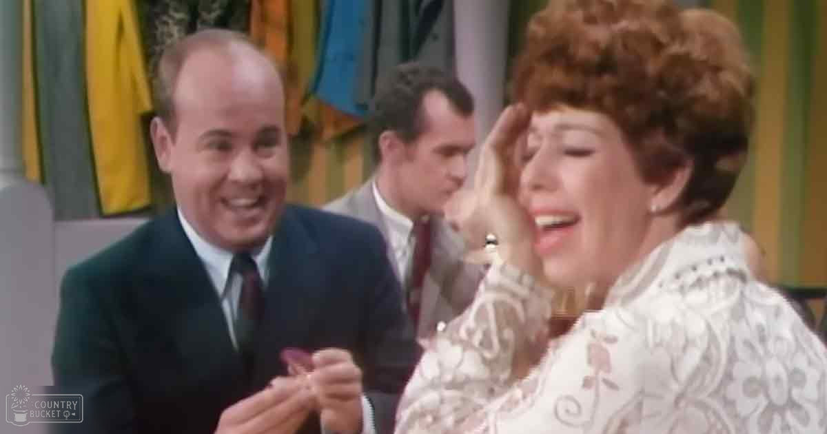 An adult female with short brown hair in a white lace-patterned blouse, surrounded by two men in suits at what appears to be an indoor social event or television setting.