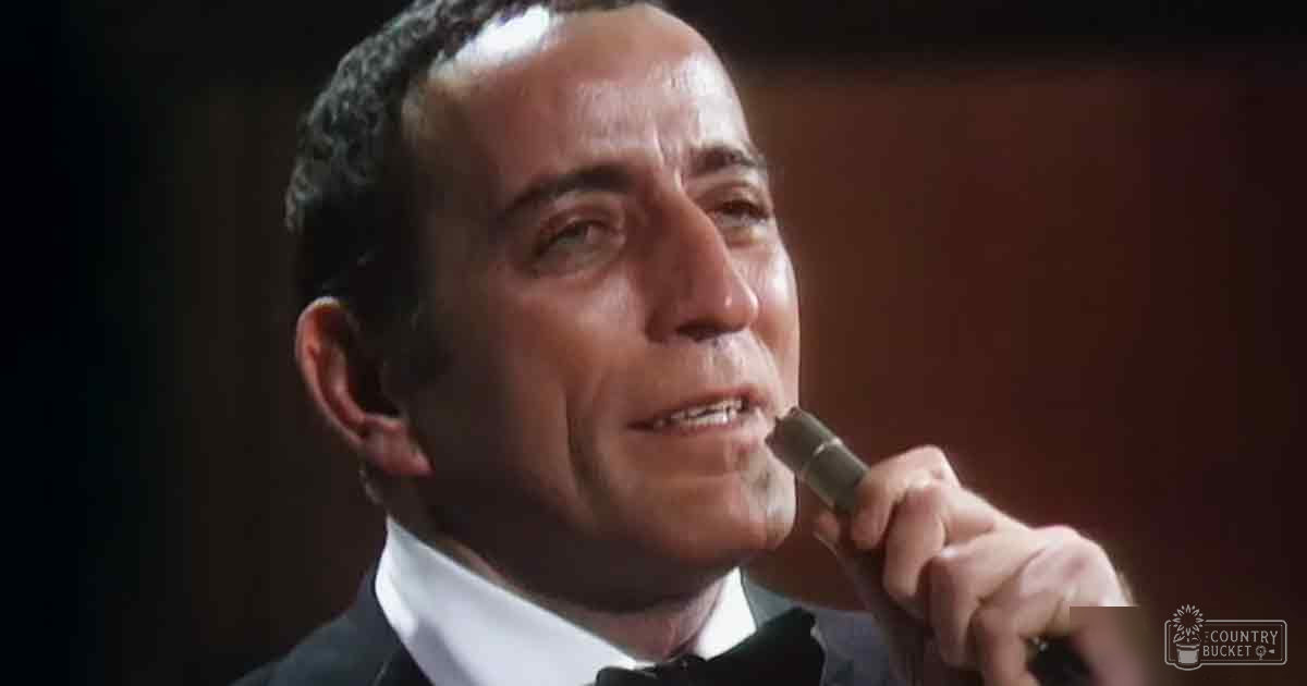 A middle-aged man in a formal black tuxedo with white dress shirt and black bow tie, holding a microphone against an indoor stage backdrop.