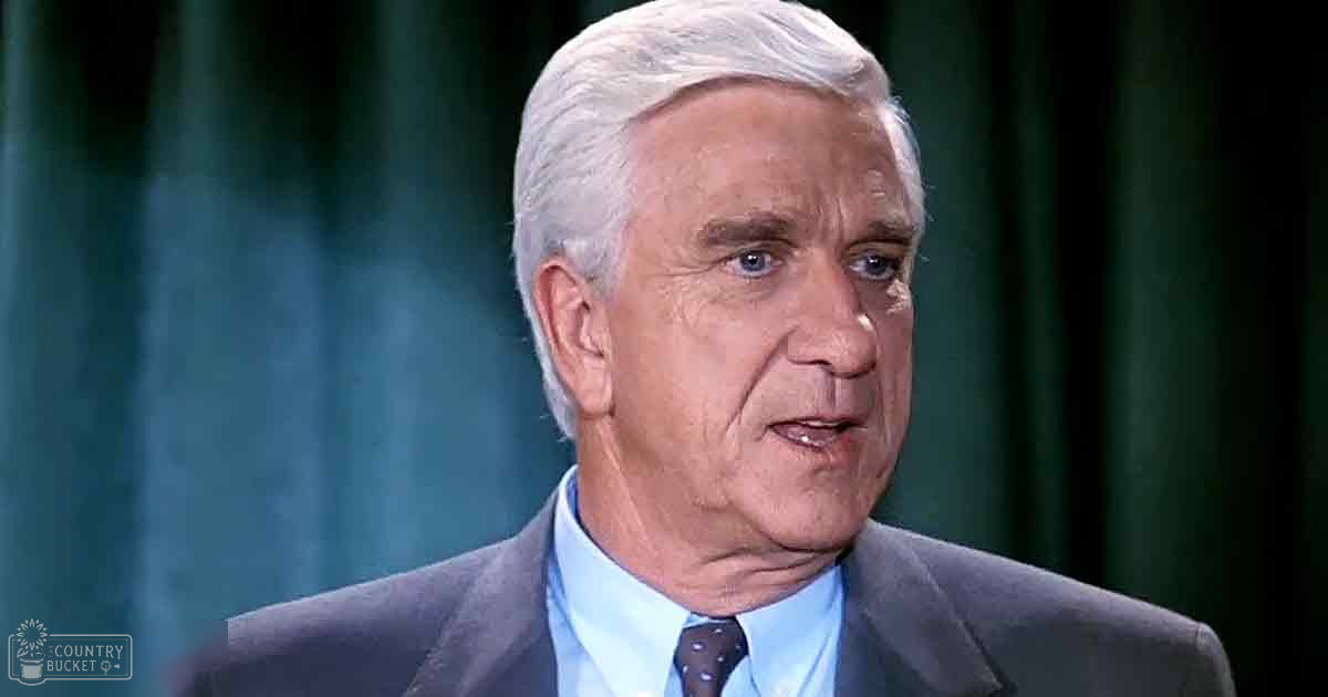 An older adult male in a tailored dark gray suit jacket and light blue collared shirt, set against a dark green curtain backdrop.