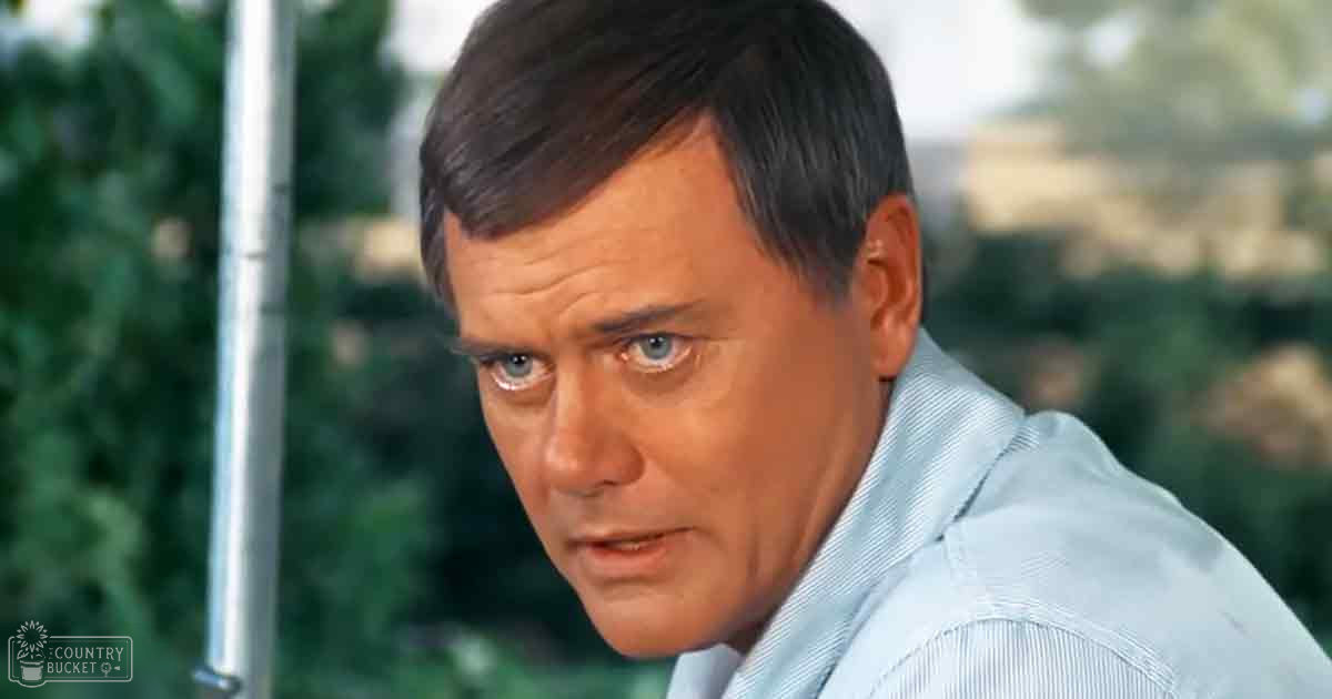 A middle-aged adult male wearing a light blue button-up shirt with vertical stripes stands outdoors amidst greenery and blurred structures.