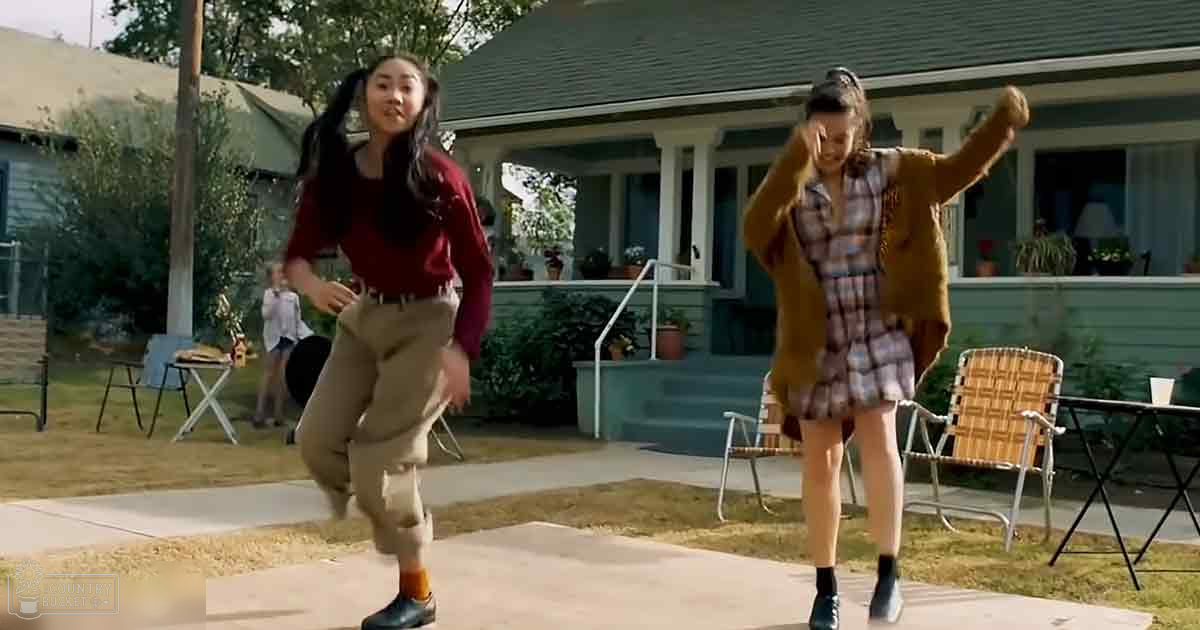 Two young adults dancing in front of a house, dressed casually in a residential neighborhood with spring or summer greenery and soft evening light.