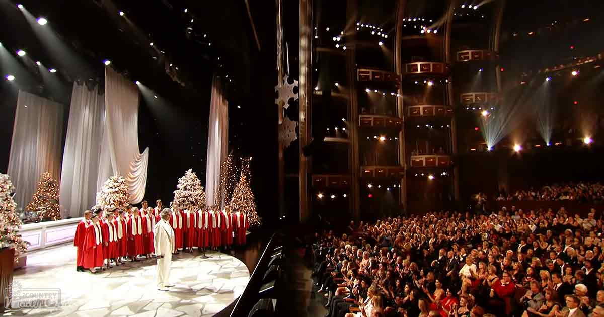 A choir performing during a holiday concert in an auditorium with Christmas trees and snowflake decorations on stage.