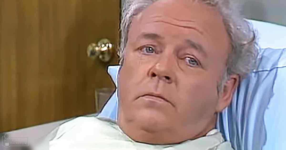 An older adult male lying in a hospital bed, wearing a light blue cotton hospital gown, with natural daylight illuminating the indoor medical setting.