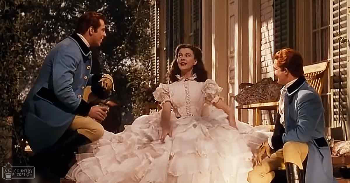 A woman in an elegant white gown sits outdoors with two men near a building with white shutters and greenery, suggesting a historical setting.