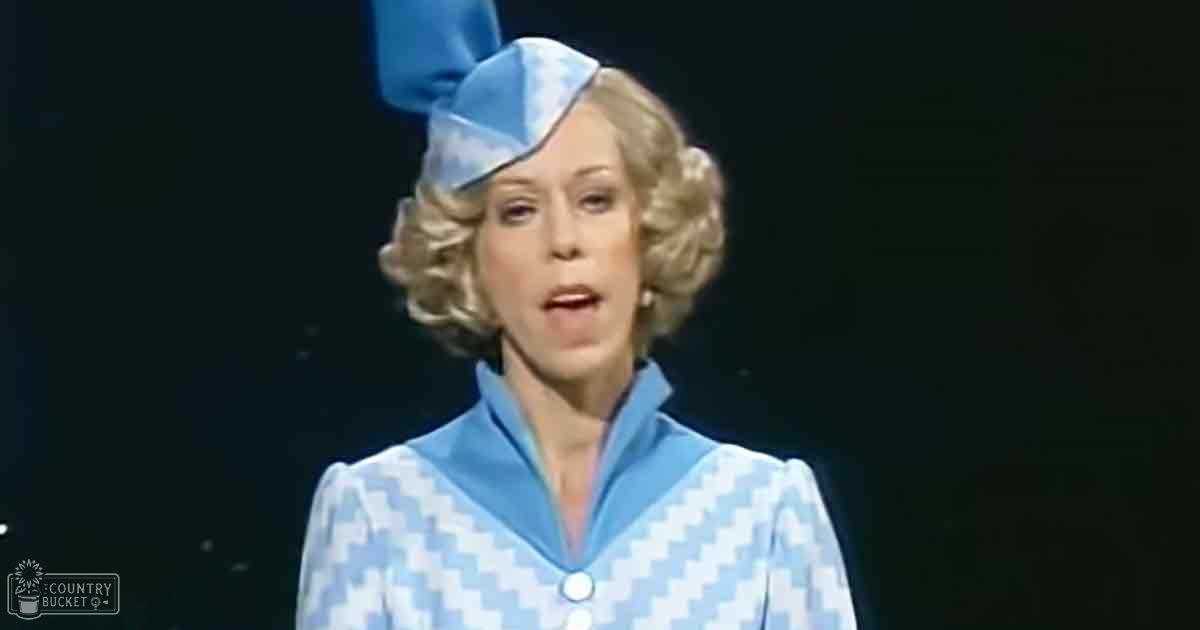 Middle-aged female wearing a blue and white checkered, collared, long-sleeved dress with a large bow on top in an indoor setting under spotlight effect.