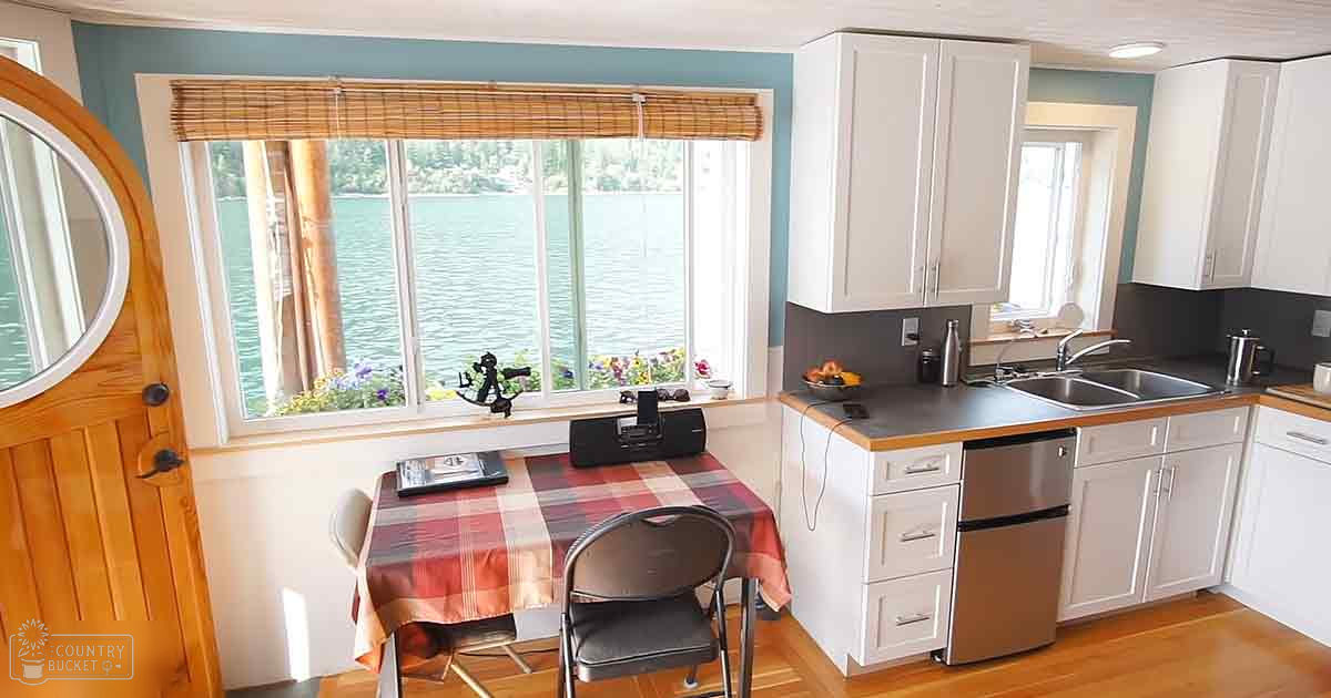 Cozy kitchen area with white cabinets, red plaid tablecloth, and a view of water through bamboo blinds in daylight near a lake or river.