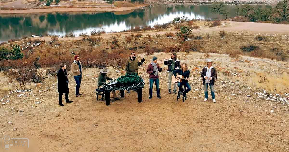 Musicians performing outdoors near a lake in an open field surrounded by dry grass and shrubs.