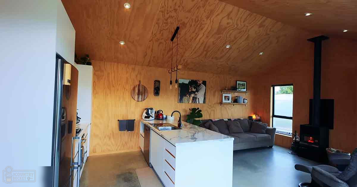A modern minimalist living space with an open-plan kitchen and living area. Wooden walls, concrete flooring, large windows allow natural light into the room.