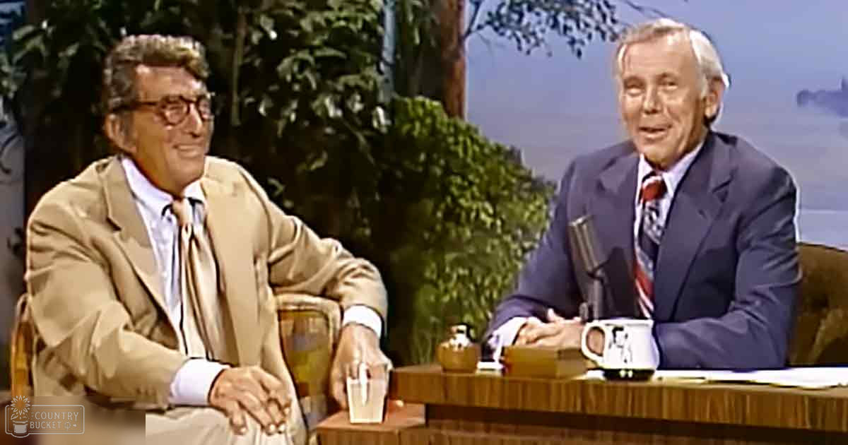Talk show host and older adult male guest seated in a studio with a backdrop of greenery and trees.