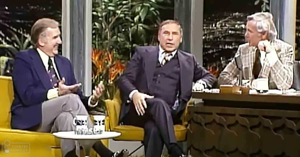 A mid-20th-century talk show set with three adults, two men and one woman, dressed in formal attire, seated against a backdrop of indoor plants and cityscape lighting.