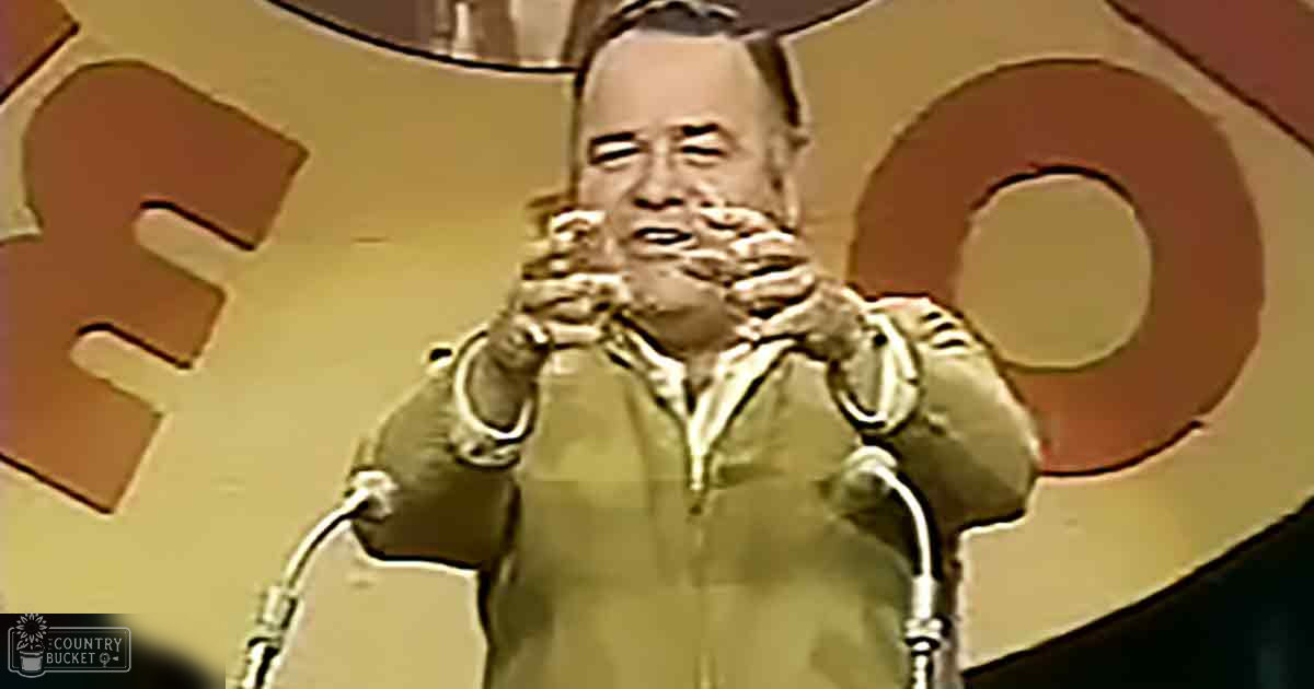 A middle-aged male performer wearing a green zip-up jacket on stage at a comedy club with professional lighting and mid-20th-century decor.