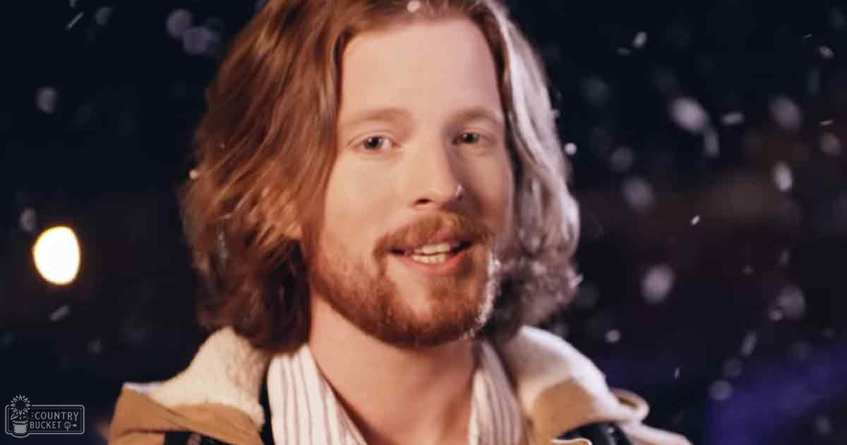 A young adult male wearing a light-colored jacket over a striped shirt with vertical lines stands indoors under soft lighting, with dark background suggesting winter evening.