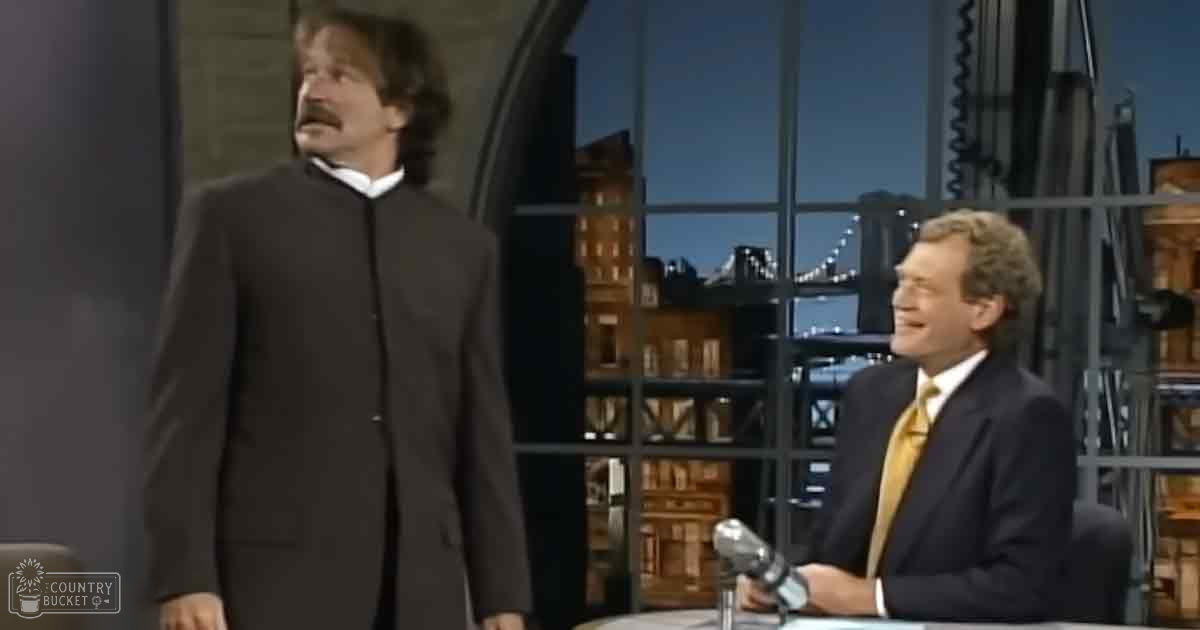 Adult male wearing a black tailored suit with a yellow tie, seated next to another adult male in a dark suit and collared shirt against an urban cityscape backdrop in what appears to be a studio setting at night.