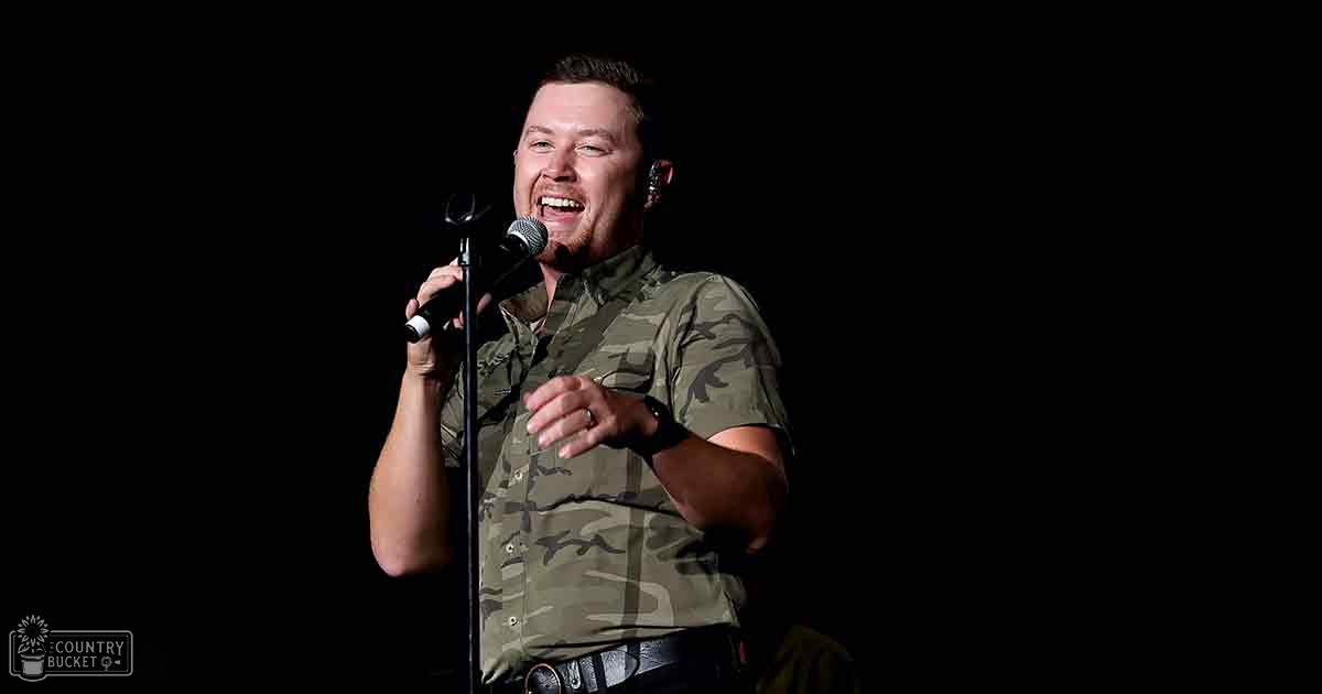 An adult male performer in a camouflage-patterned short-sleeve shirt with a button-down front, standing indoors near a microphone stand and wearing a black wristband.