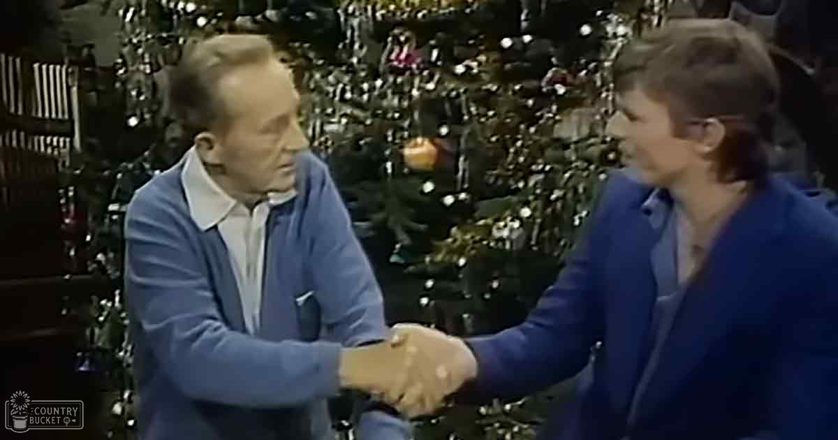 Two middle-aged men shaking hands in front of a decorated Christmas tree, suggesting an indoor winter holiday setting with warm lighting and festive decor.