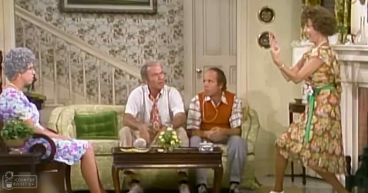 A woman wearing a floral dress with short sleeves, holding an accessory, surrounded by two men in casual attire and another woman in similar dress, indoors in a living room setting.
