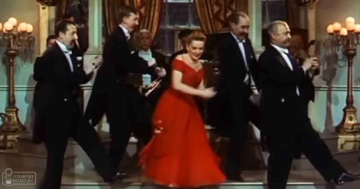 An adult female dancer in a red ball gown dancing with men in tuxedos at an elegant indoor event.