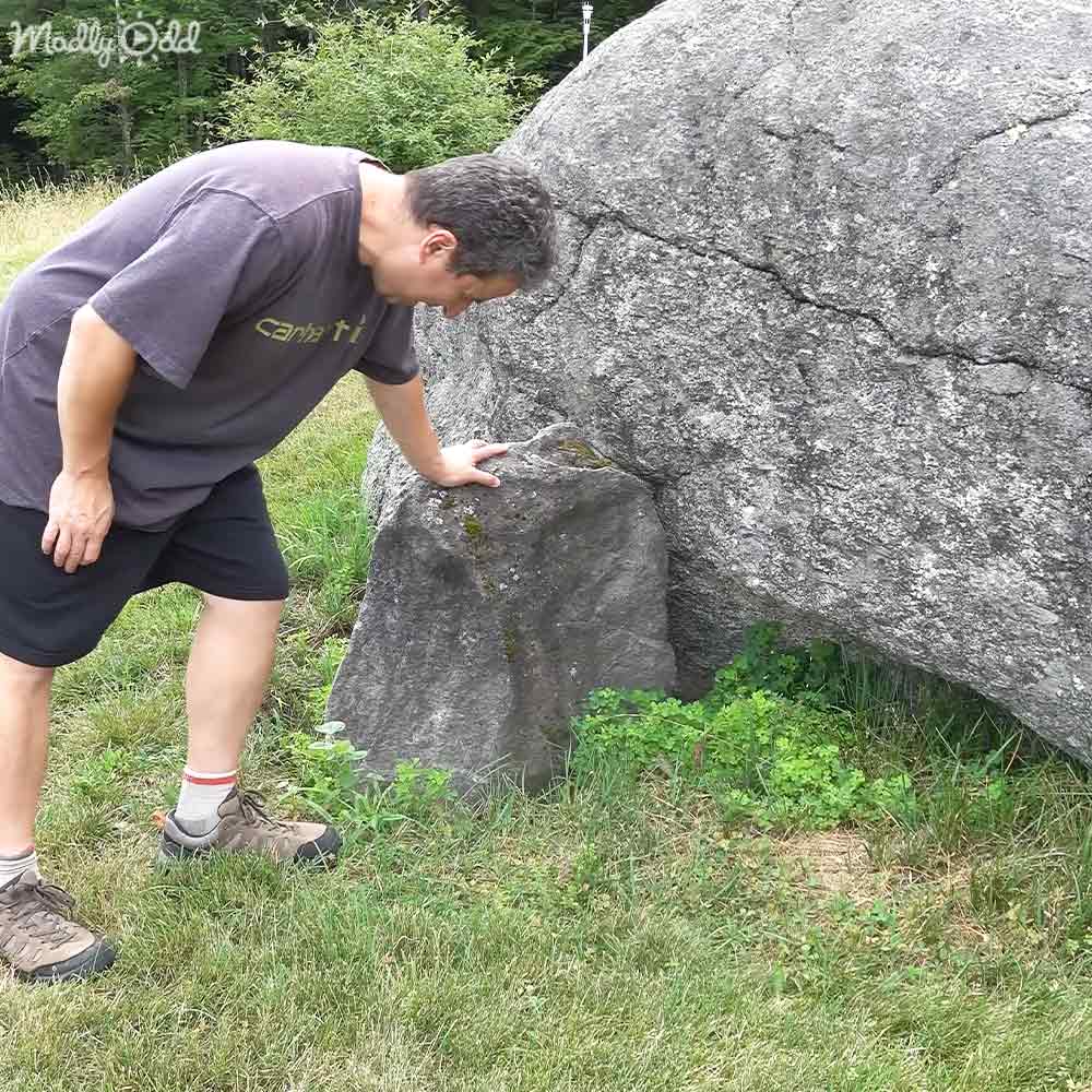 An adult male in a loose-fitting dark gray t-shirt and brown hiking boots, interacting closely with a large rock in an outdoor park setting during spring or summer under natural daylight.