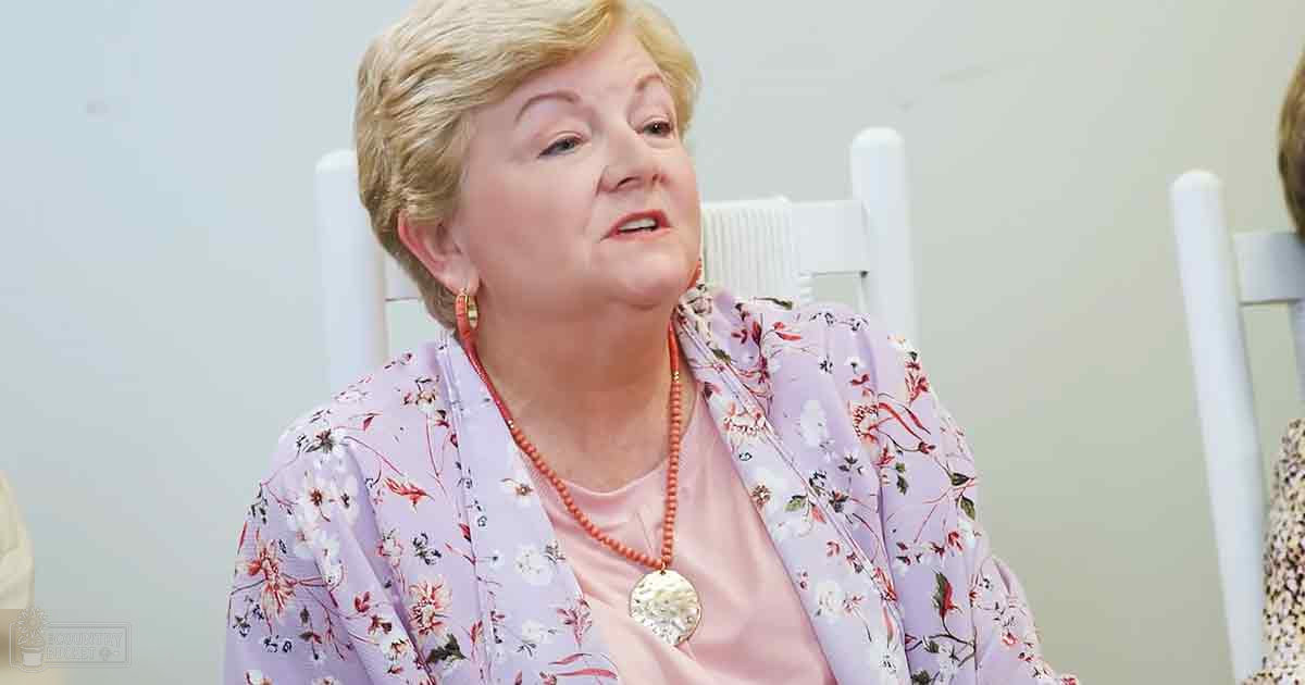 An older adult female wearing a light purple floral-patterned blouse and long necklace with orange beads, casual indoor setting with white chairs and minimal decor.