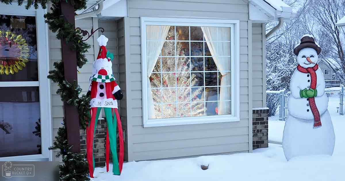 Festive winter scene with a large snowman decoration wearing a red scarf and hat in front of a house, adorned with Christmas decorations visible through the window.