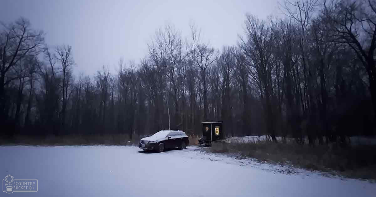 A snowy rural landscape with a dark-colored SUV parked near a small wooden structure in dense trees, under an overcast sky.
