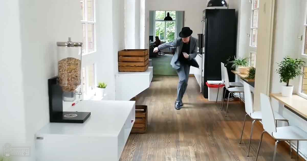 A young adult male wearing a gray blazer, black shirt, dark gray trousers, dark shoes, and a black fedora hat in an office or co-working space with white walls, brown wooden crates, white chairs, plants, and large windows allowing natural light to enter.