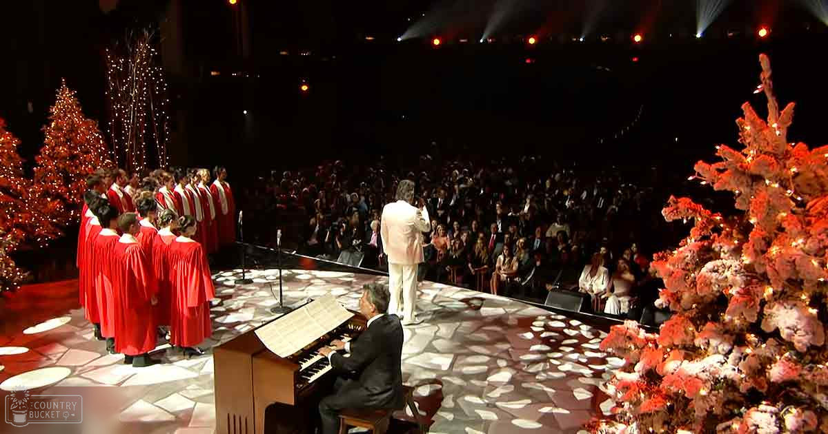 A Christmas concert in an indoor auditorium with a choir dressed in red robes performing on stage under artificial lighting, surrounded by decorated Christmas trees.