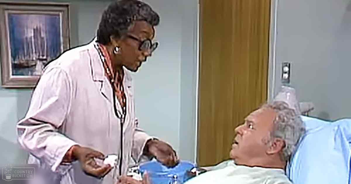 A healthcare professional in a hospital room with an older adult male patient wearing a light blue hospital gown. The setting includes a hospital bed, wooden door, and framed picture on the wall.