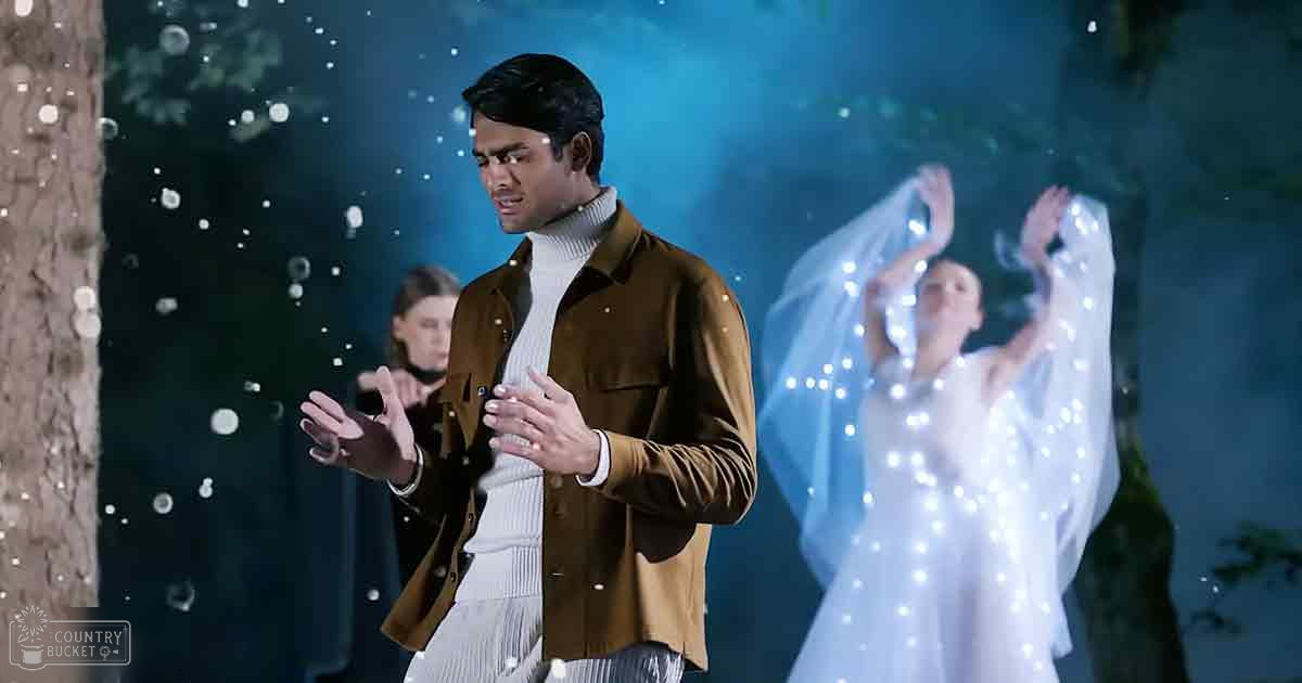 An adult male in a brown jacket and white turtleneck sweater stands against an ethereal backdrop with snowflakes falling, while behind him, an adult female in a white dress adorned with small lights holds a veil or fabric that extends upwards. The scene is set outdoors at night under a dark blue sky dotted with glowing particles, creating a magical or dreamlike atmosphere.