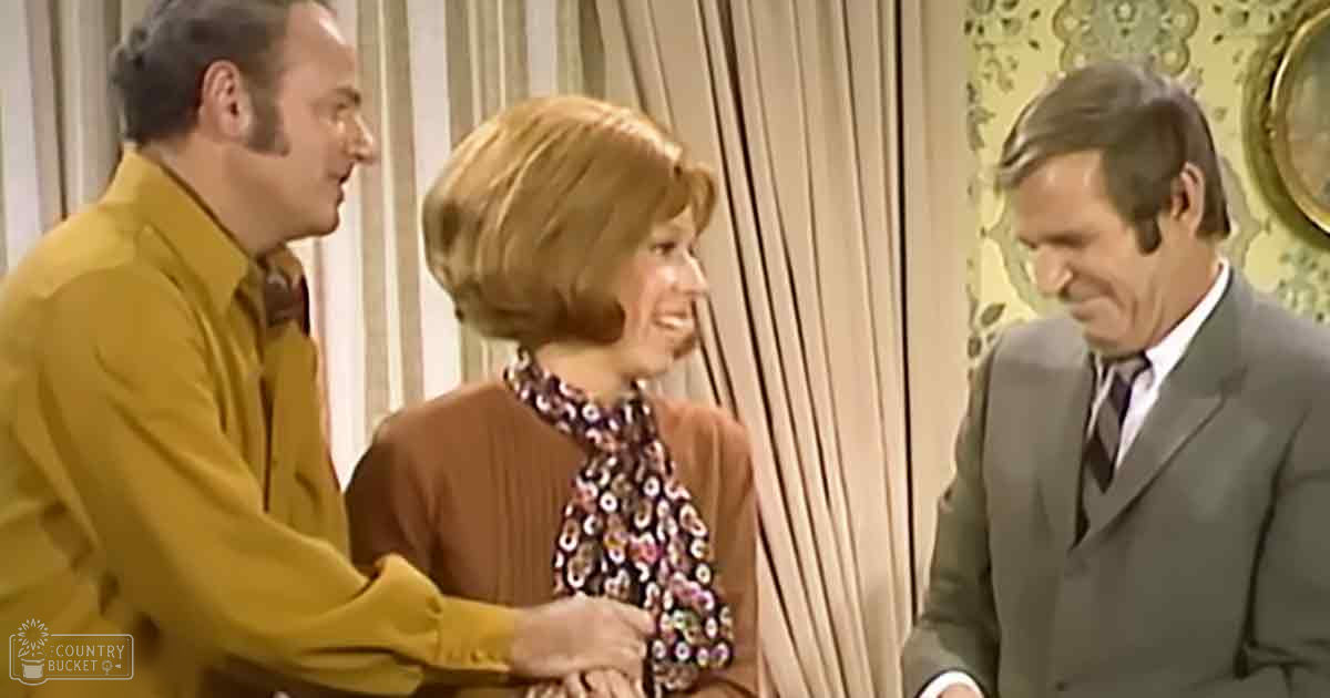 Adult male in mustard yellow blazer with patterned shirt; adult female in brown cardigan over floral blouse; adult male in gray suit jacket over white dress shirt, all indoors with striped curtains and a circular gold-framed mirror. Likely mid-20th century setting.