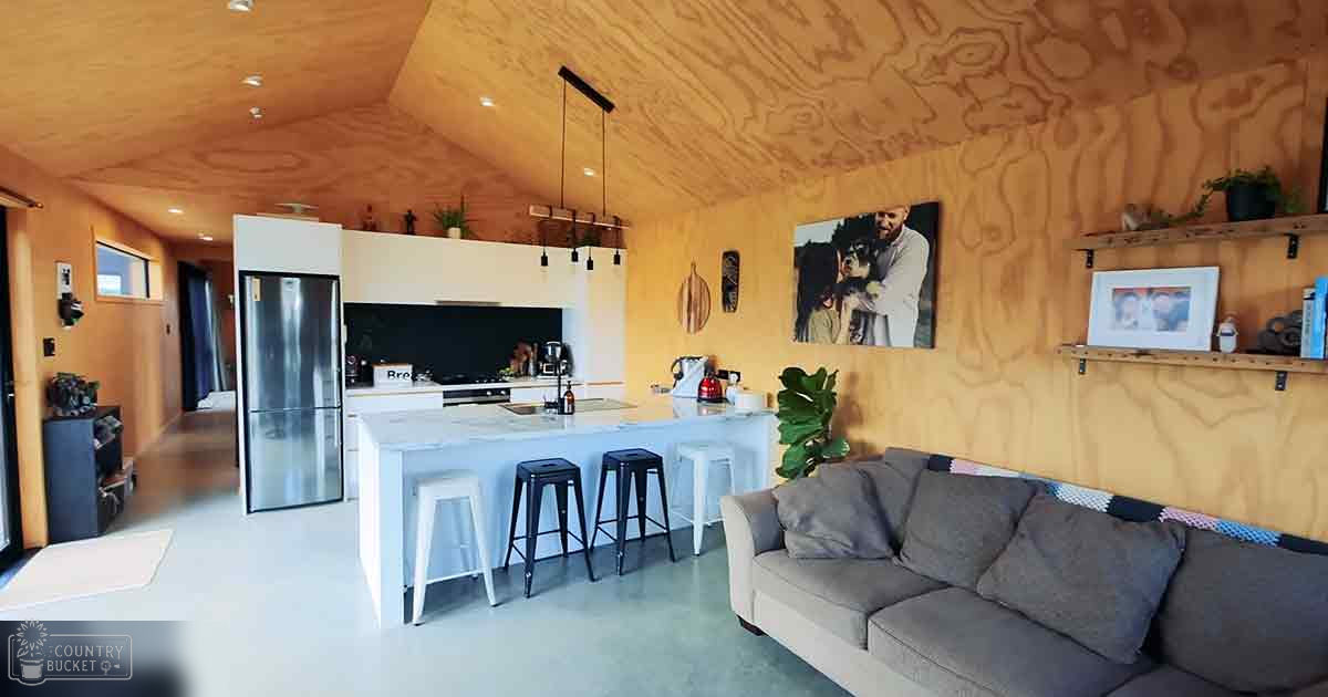 A modern home interior with an open kitchen area with a gray sofa, white island, wooden shelves holding plants and photos, and natural light from the day.