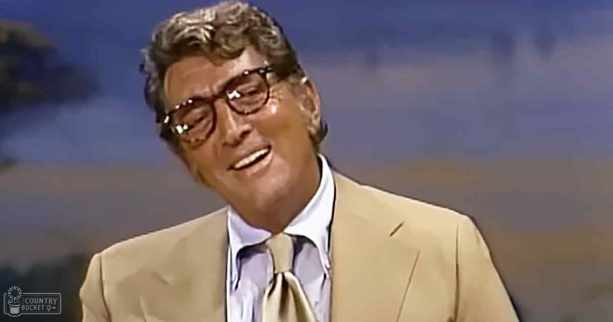 An older adult male wearing a light-colored suit jacket and a collared shirt with glasses in a studio setting with blurred mountains in the background, suggesting daytime lighting.