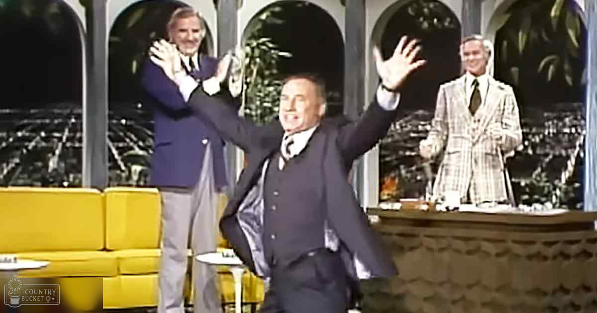 An adult male performer on stage in a dark suit and white shirt, holding a cane; older adults wearing similar suits, clapping hands; mid-century entertainment setting with arched windows, yellow sofa, round table, bar counter, decorative elements.
