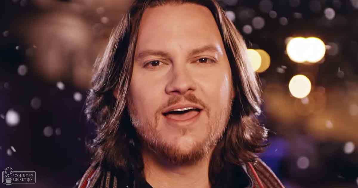 An adult male wearing a dark, patterned shirt in an indoor setting with warm bokeh lighting suggesting a festive or celebratory atmosphere.