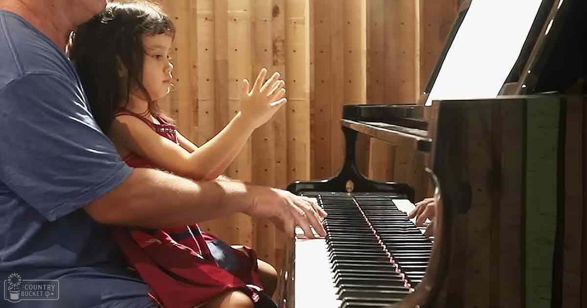 A toddler wearing a red floral or abstract dress, learning to play the grand piano with an adult in a cozy home setting under natural sunlight.