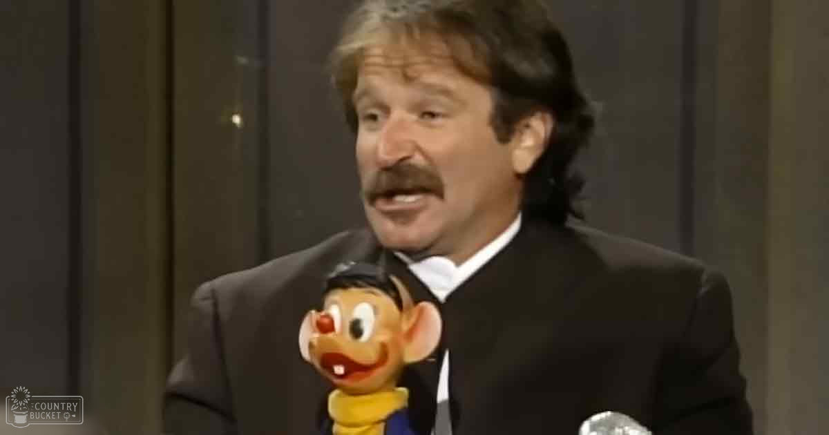 Middle-aged male in a well-fitted dark suit jacket over a white shirt and black tie, holding a Goofy puppet resembling Disney's character, within an indoor studio setting with dark curtains suggesting daytime or artificial lighting.