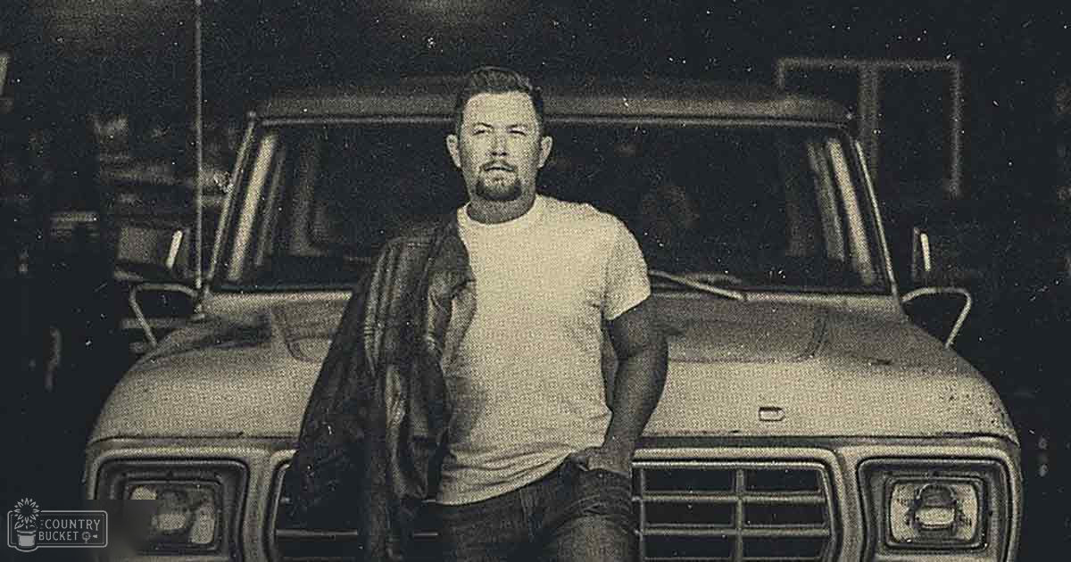 A middle-aged man wearing a plain white t-shirt stands next to a vintage pickup truck at night, with artificial lighting illuminating the scene.