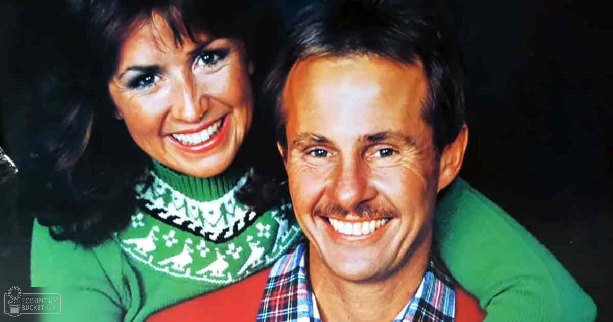 A couple indoors with a female wearing a green turtleneck sweater and male in a red plaid shirt under a green jacket, set against dark evening lighting.
