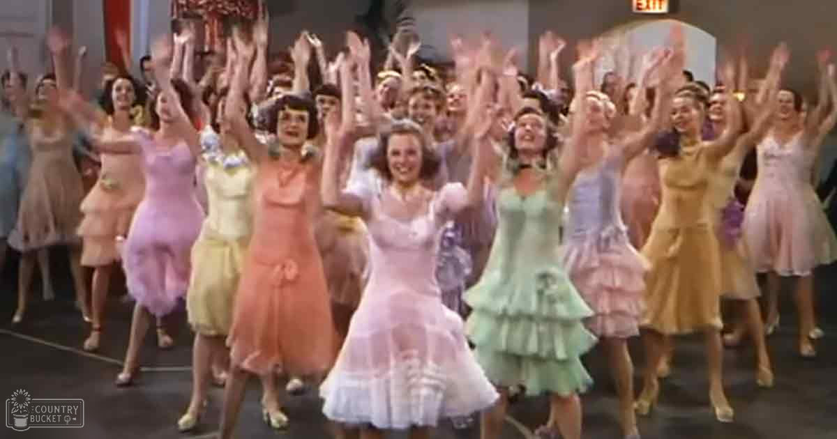 A mid-20th century dance scene with young adult female dancers on a stage, wearing pastel-colored dresses typical for the era.